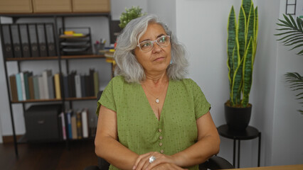 Grey-haired woman sitting in an office room with bookshelves and plants in the background.