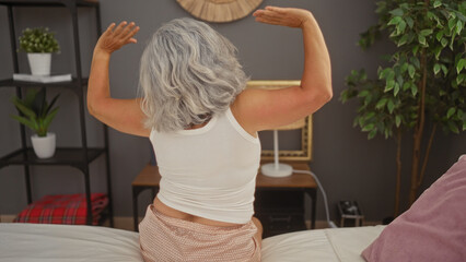 Mature grey-haired woman stretching in bedroom at home with indoor plants and decor in the background