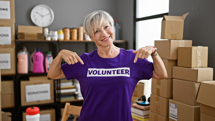 A smiling, mature woman wearing a volunteer shirt points at herself in a warehouse full of donation...