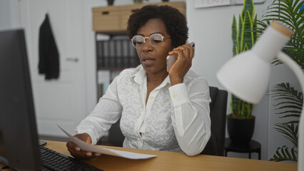 Middle-aged african american woman with curly hair and glasses working in a modern office, talking on the phone and reviewing documents.