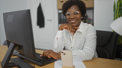 Woman smiling at office desk with computer, indoor workspace, curly hair, glasses, african american, professional attire, middle-aged, working environment, white shirt