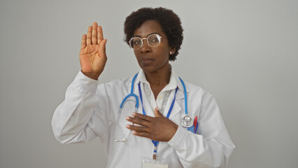 Female doctor standing against a white background with her hand raised, taking an oath or making a pledge with a serious expression