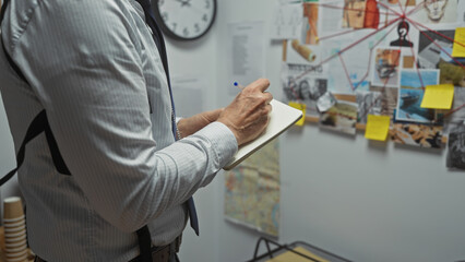 A mature man takes notes in a police station with a clue board full of photos and strings