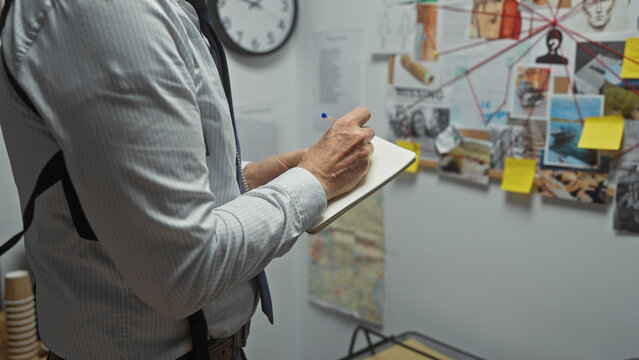 A mature man takes notes in a police station with a clue board full of photos and strings