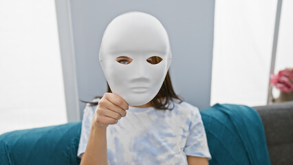 A young woman holds a blank white mask in a modern living room, suggesting anonymity or mystery.