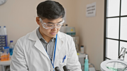 Young asian man working in a laboratory, wearing safety goggles and a lab coat, surrounded by scientific equipment.