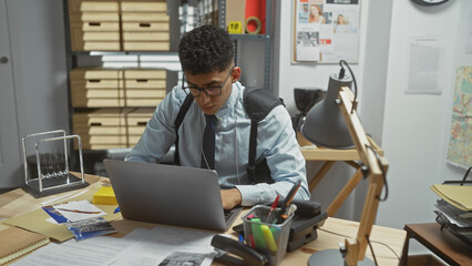 A concentrated young man working on a laptop in a cluttered detective's office filled with files...