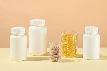 Healthcare product theme photo, was taken from frontal against beige background. Two glass beaker contain vitamin capsules displayed in center, white unlabeled medicine bottle placed on both sides