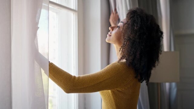 Camera view of young woman with curly hair. African American girl standing by window. Opening curtains and looking out into street with slight smile on her face. Natural lighting. Calm atmosphere.