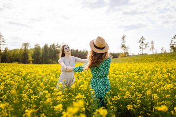 Fototapeta premium Two young women in a beautiful field with yellow flowers. Summer day walks in flower plantings. Girlfriends together.