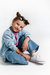 Cute kid girl model with buns wearing sunglasses, jeans and pink t-shirt sitting and posing on white studio background