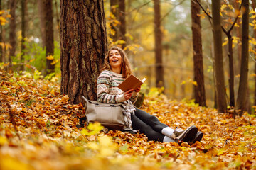 Cute woman reads a book sitting in a autumn yellow park under a tree. Education. Concept of enjoying nature.