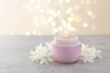 Cream in jar and beautiful jasmine flowers on grey table against light background with blurred lights, closeup
