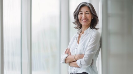 An elegant woman in a white blouse stands confidently in a bright, modern office with her arms crossed, smiling warmly at the camera.