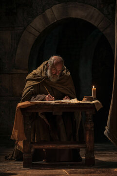 A senior prophet disciple in a candle lit room or dungeon writing the scriptures. Ancient medieval man writing on his desk. Bald and beard. Worn robe. In a stone room. Religious concept art.