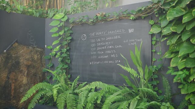 Chalkboard with Hurty Hundred workout plan at upscale outdoor gym in tropical setting, surrounded by greenery, gym equipment. Fitness enthusiasts follow intense exercise routine in retreat atmosphere.