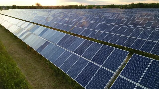 Aerial flyover footage of solar panel photovoltaic energy farm in a green farmland field. High angle shot of pv cells array ssytem generating electricity on a rural summer afternoon. Renewable concept