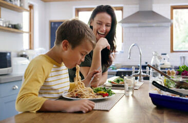 Happy woman, child and eating with spaghetti at kitchen table for meal, food and nutrition or healthy diet. People, excited and hungry for family dinner, weekend and bonding together in home with joy