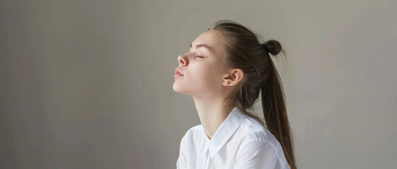 Fototapeta premium A serene young woman with closed eyes, appearing relaxed and meditative, wearing a crisp white shirt against a minimalistic background.