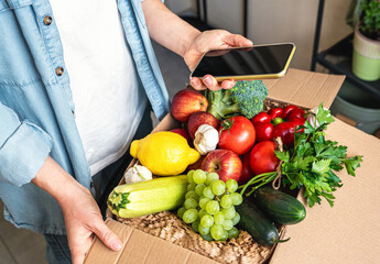 Online ordering farm grown vegetables and fruits. Mobile phone with blank screen in hand of female person and cardboard box with fresh vegetables.