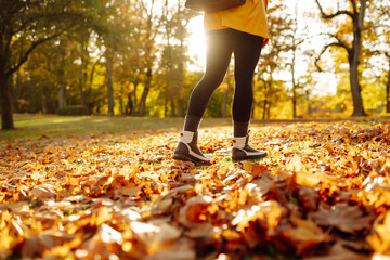 Close-up of female legs in hiking boots walks on ground with yellow-orange dry fall leaves during autumn season in park or forest. Feet walking in outdoor nature. Healthy lifestyle on leisure activity