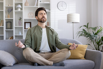 Man meditating on sofa in living room, practicing mindfulness and achieving relaxation. Person...
