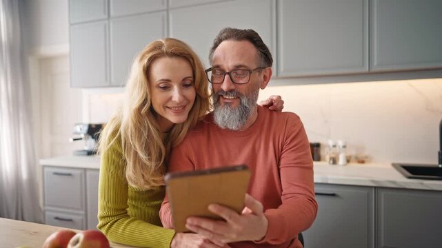 Older Caucasian male holding very small tablet device or mobile phone. Using glasses for better vision. Showing screen of gadget to beloved wife with long blond hair. Discussing something together.