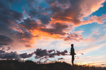 Silhouette of a lonely man in the green grass of a hill against the background of a sunset cloudy sky. The figure of a man against the sky.