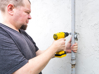 A man uses a cordless drill to secure a metal pole to a white wall. He is wearing a gray t-shirt...