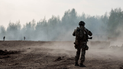 A soldier in camouflage and ammunition stands on a dusty road, a blurred forest in the background and figures of other military troops.