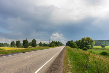 Fototapeta premium A tranquil road meanders through vibrant greenery as dark clouds loom above, hinting at an impending change in weather.