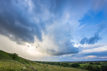 Dark clouds gather and shift in the evening sky, casting a dramatic shadow over the rolling green...