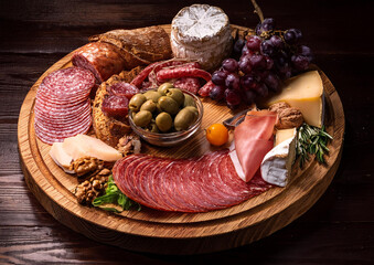 Still life scene. A circular wooden tray with some delicatessen such as olives, cold cuts, cold meat, salami, cheese and grapes