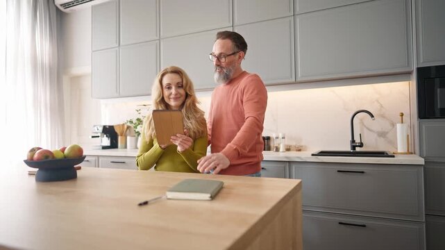 Warm moment of Caucasian couple. Man standing besides his wife. Woman giving fresh apple to beloved husband. People talking with each other. Female using large phone or small tablet device.