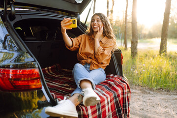 Beautiful woman is resting in trunk of car and taking selfie. Travel, weekend, nature,  relax and lifestyle concept