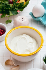 Greek yogurt in a white bowl with a yellow rim on a white table with ingredients laid out around, salt shaker, lemon, herbs, garlic