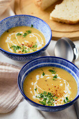 Two bowls of creamy potato soup, spoons, napkin and wooden cutting board with a loaf of bread and cut pieces. The surface of the soup is decorated with cream, croutons, parsley and ground red pepper.