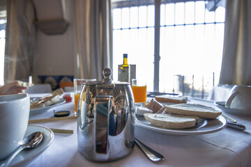 A table with the typical breakfast, chrome coffee maker, cutlery, cups, juices, bread, olive oil. In the background a large window with lots of natural light.