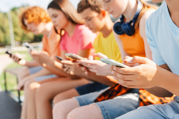 Cheerful children using mobile phones sitting in a row on bench in the city