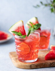 Two glasses watermelon cocktail with lime, ice and mint on a wooden board on a light background with branch and fruits close up.