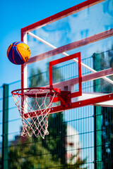 A yellow-blue basketball ball flies into a red hoop with a net against a blue sky