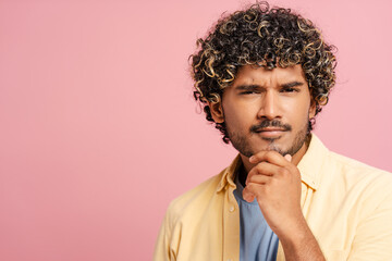 Fototapeta premium Pensive young Indian adult man touching chin with hand on pink background