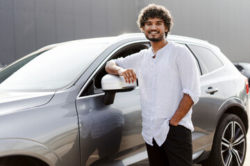 Handsome, indian young man is leaning on his new grey car smiling at the camera
