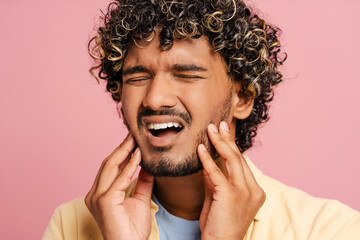 Young Indian man suffering from strong toothache touching cheek on pink background