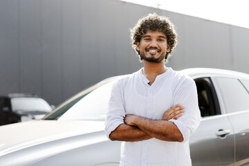 Confident indian man smiling with arms crossed in front of a new car