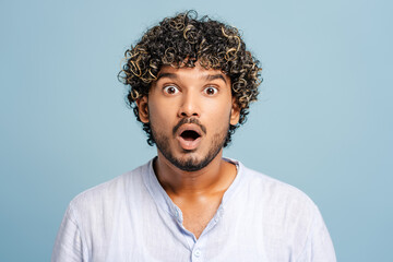 Shocked Indian young man posing on blue background, showing surprise emotion, opening mouth widely