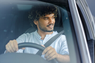 Attractive, smiling Indian man driving a car, steering wheel, looking away. New transportation