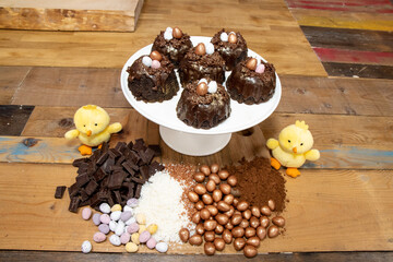 A selection of Easter Chocolate nest Cakes topped with three little chocolate mini eggs on a white plate surrounded by the ingredients and two yellow chicks
