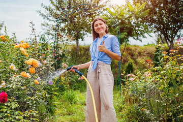 Woman gardener watering plants with hose pipe in summer garden showing thumb up. Taking care of...