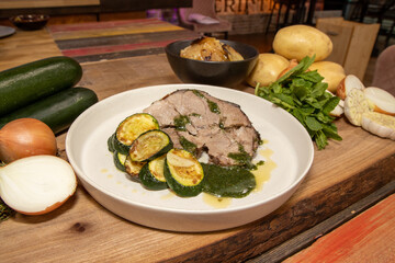 A delicious plate of Slow Roasted Lamb and Boulanger Potatoes on a wooden kitchen table surrounded by the ingredients used to create the meal